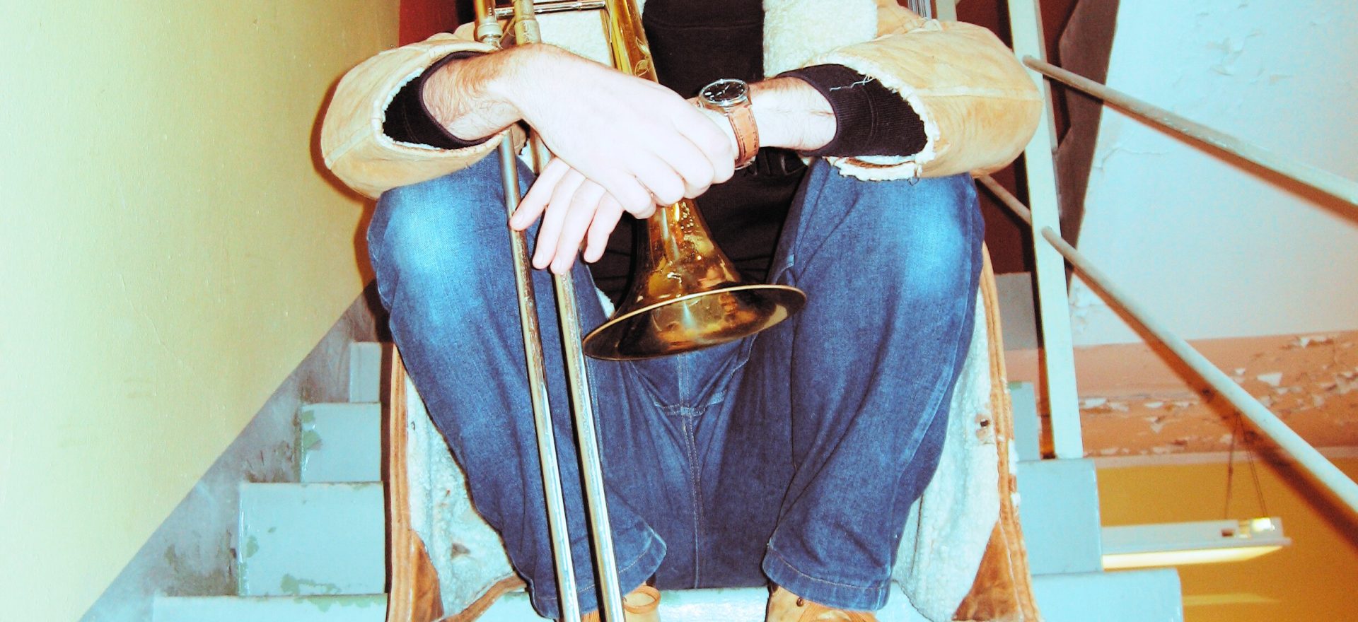 Mann sit i trappeoppgang, ser mot eine sida. Kort hår, bart. Held ein trombone framføre seg.