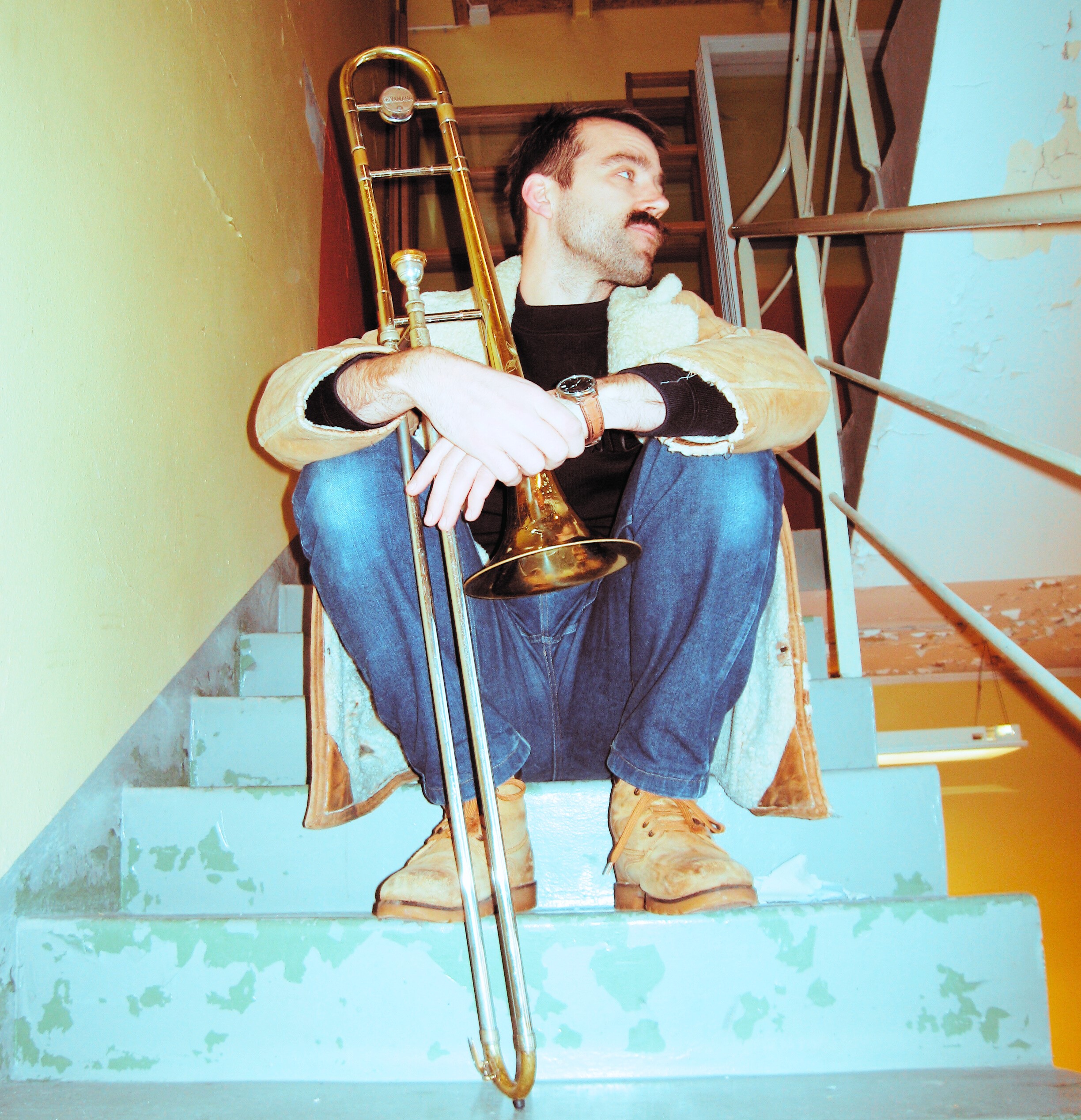 Mann sit i trappeoppgang, ser mot eine sida. Kort hår, bart. Held ein trombone framføre seg.