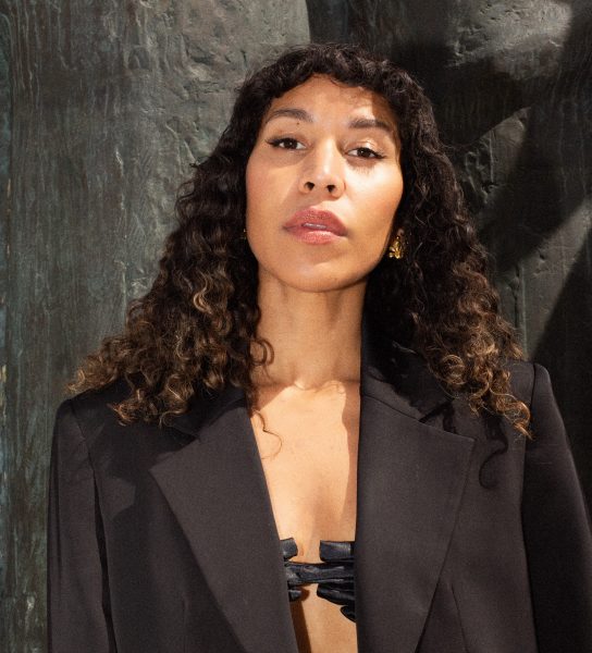 Kvinne ser rett i kamera. Svart dressjakke, open. Mørke krøllar ligg mjukt på skuldrene, pannelugg. Mørk bakgrunn, steinvegg.