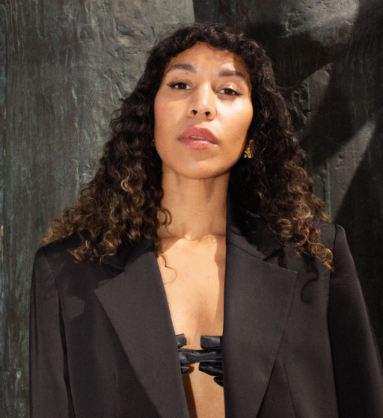 Kvinne ser rett i kamera. Svart dressjakke, open. Mørke krøllar ligg mjukt på skuldrene, pannelugg. Mørk bakgrunn, steinvegg.