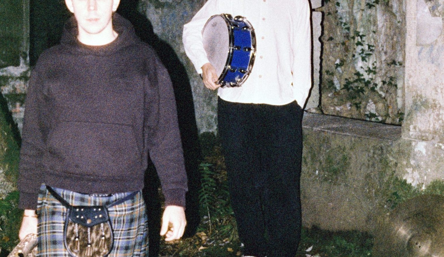 To menn er ute i mørket, opplyst med stertkt lys. Mann til venstre har kort hår, svart hettegenser, kilt, knestrømper, tradisjonell skotsk veske og har ein saxofon i handa. Mannen til høgre har hue, kvit skjorte med mandarinkrage, svart bukse, kvite sneakers, og han held ei knallblå skarptromme under eine armen. Dei ser ut til å stå framfor eit gamalt ruinbygg.