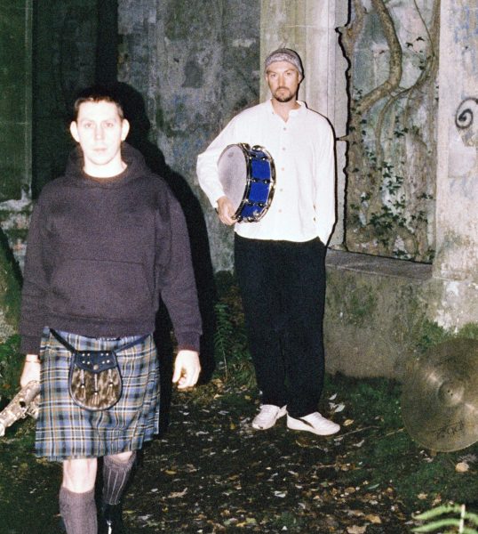To menn er ute i mørket, opplyst med stertkt lys. Mann til venstre har kort hår, svart hettegenser, kilt, knestrømper, tradisjonell skotsk veske og har ein saxofon i handa. Mannen til høgre har hue, kvit skjorte med mandarinkrage, svart bukse, kvite sneakers, og han held ei knallblå skarptromme under eine armen. Dei ser ut til å stå framfor eit gamalt ruinbygg.