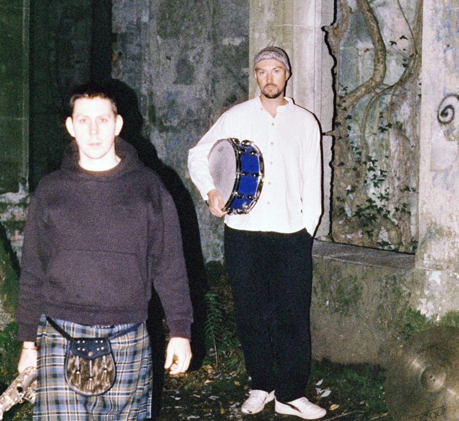 To menn er ute i mørket, opplyst med stertkt lys. Mann til venstre har kort hår, svart hettegenser, kilt, knestrømper, tradisjonell skotsk veske og har ein saxofon i handa. Mannen til høgre har hue, kvit skjorte med mandarinkrage, svart bukse, kvite sneakers, og han held ei knallblå skarptromme under eine armen. Dei ser ut til å stå framfor eit gamalt ruinbygg.