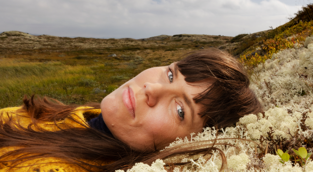 Benedicte Maurseth ligg på ryggen og kviler hovudet i lys lyng, ho ser mot kamera. Brunt hår, pannelugg. Hardangervidda i bakgrunnen.